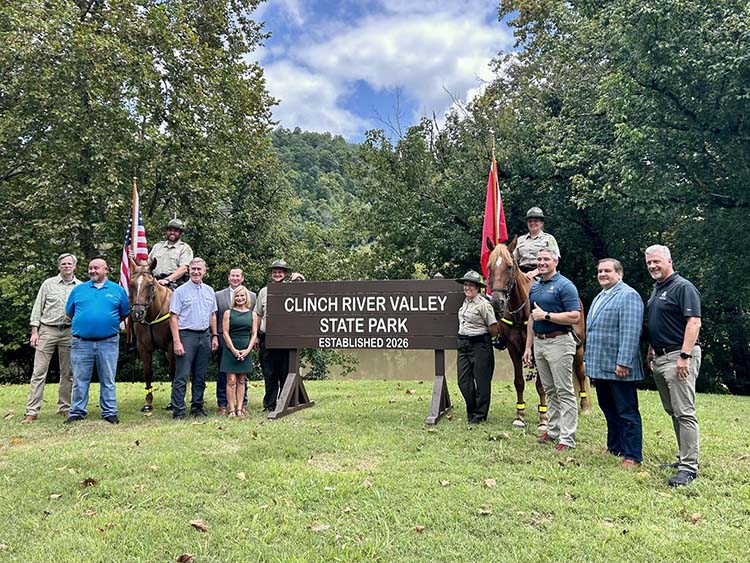 A group of people, including uniformed state park rangers and two horses, are positioned around a large wooden sign that reads